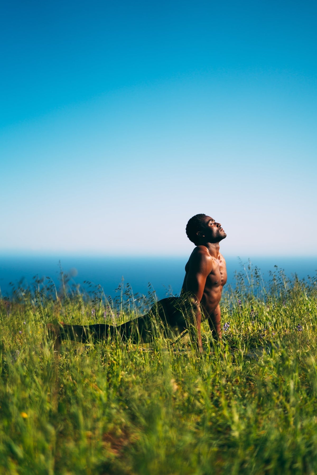 Man performing Upward Dog while in tall grass overlooking the ocean.