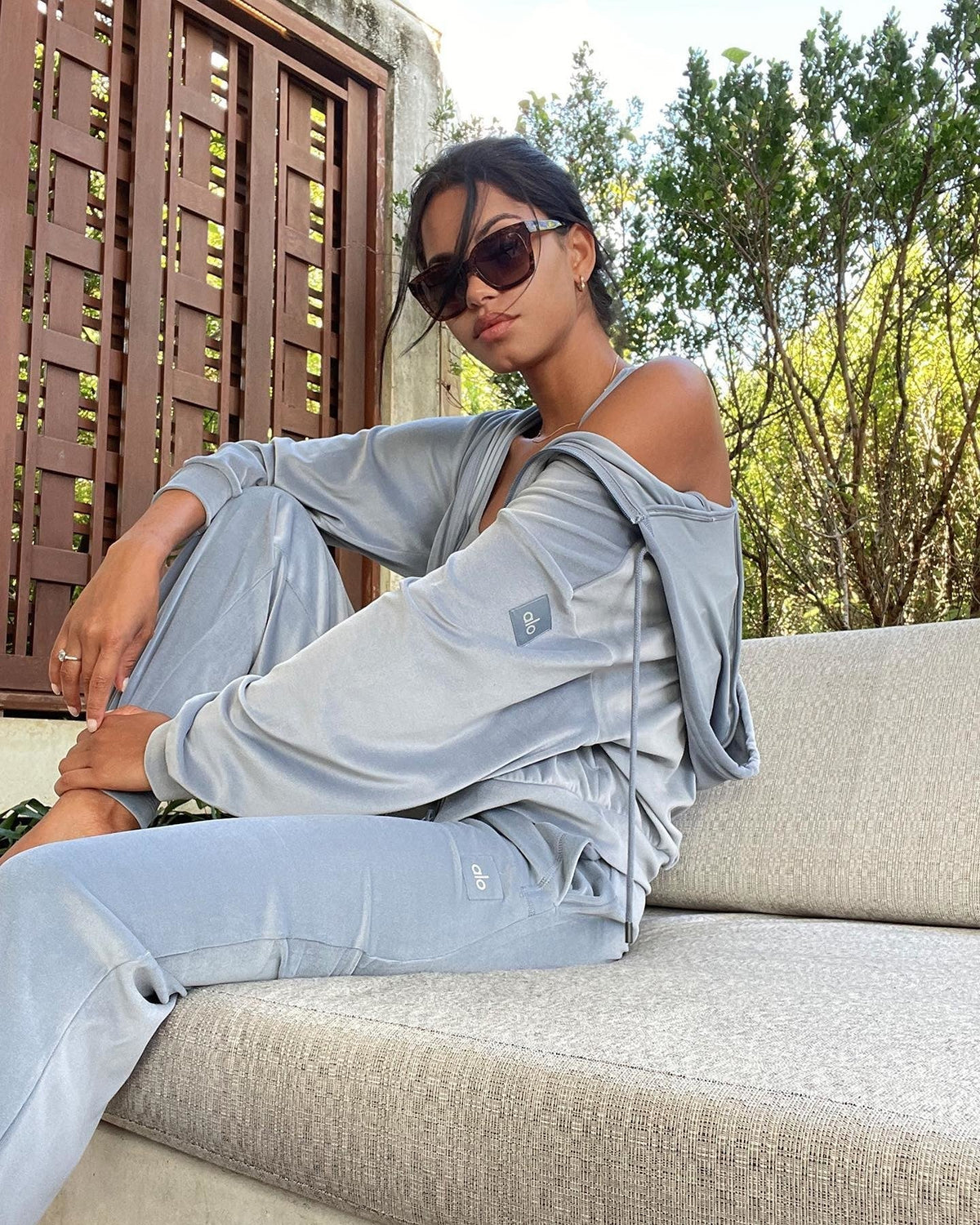 @daianesodre wearing the Velour Glimmer Full Zip Hoodie and Velour High-Waist Glimmer Wide Leg Pant in Steel Blue while seated on a couch posing for the camera.