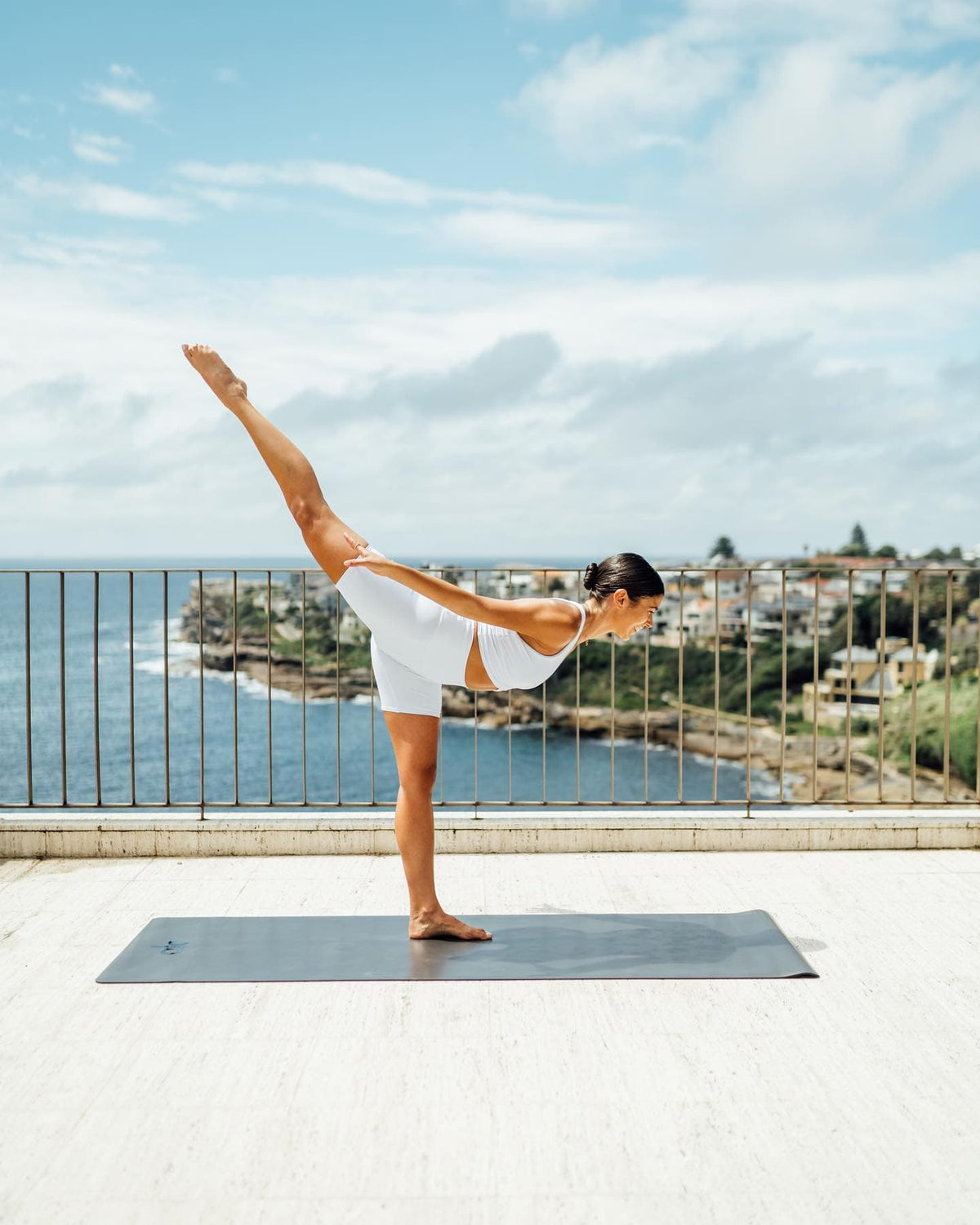 @biancamelas in warrior III pose on an Alo Warrior Mat atop a balcony overlooking the ocean.