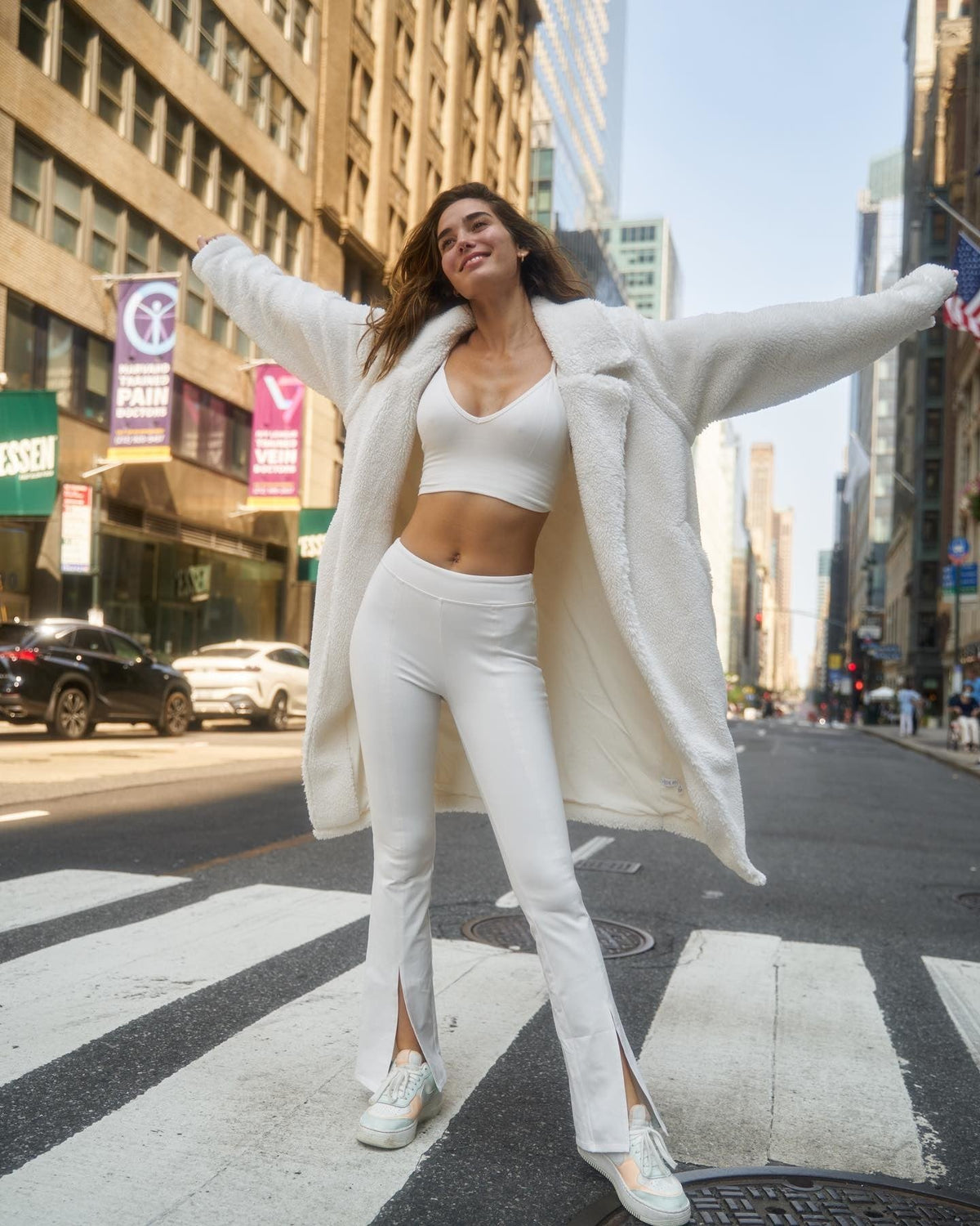 @rachellvallori wearing White High-Waist Flutter Leggings with a White Delight Bralette, a White Sherpa Trench and white sneakers while posing in a crosswalk in a big city.